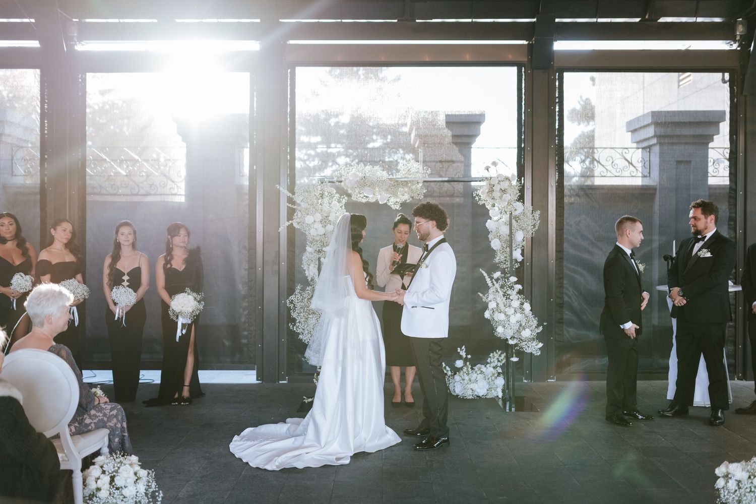A sunlit wedding ceremony space with white floral arrangements and guests looking on.