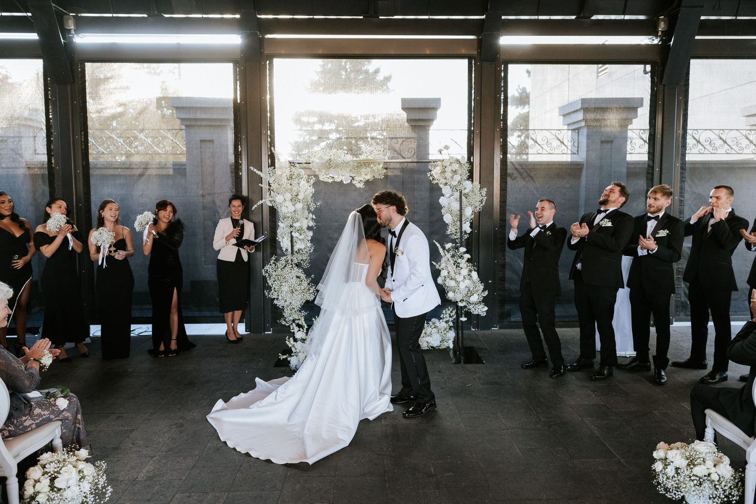 A wedding couple shares a romantic moment as guests applaud in front of floor-to-ceiling windows.