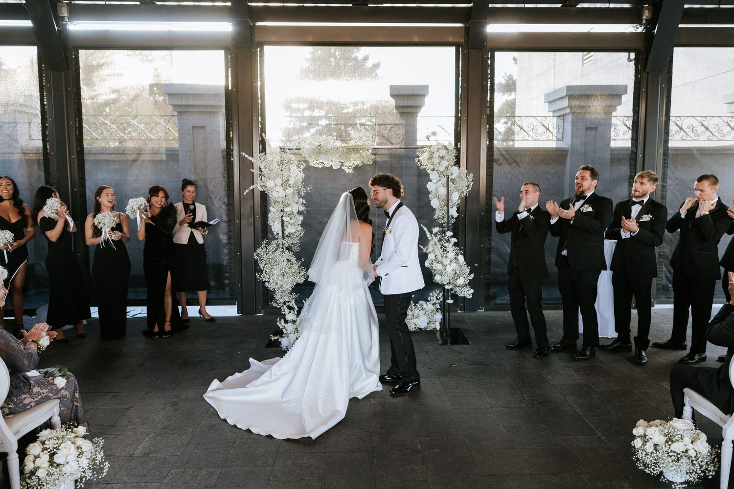 Modern wedding ceremony with guests watching couple at indoor venue decorated with white floral arrangements.