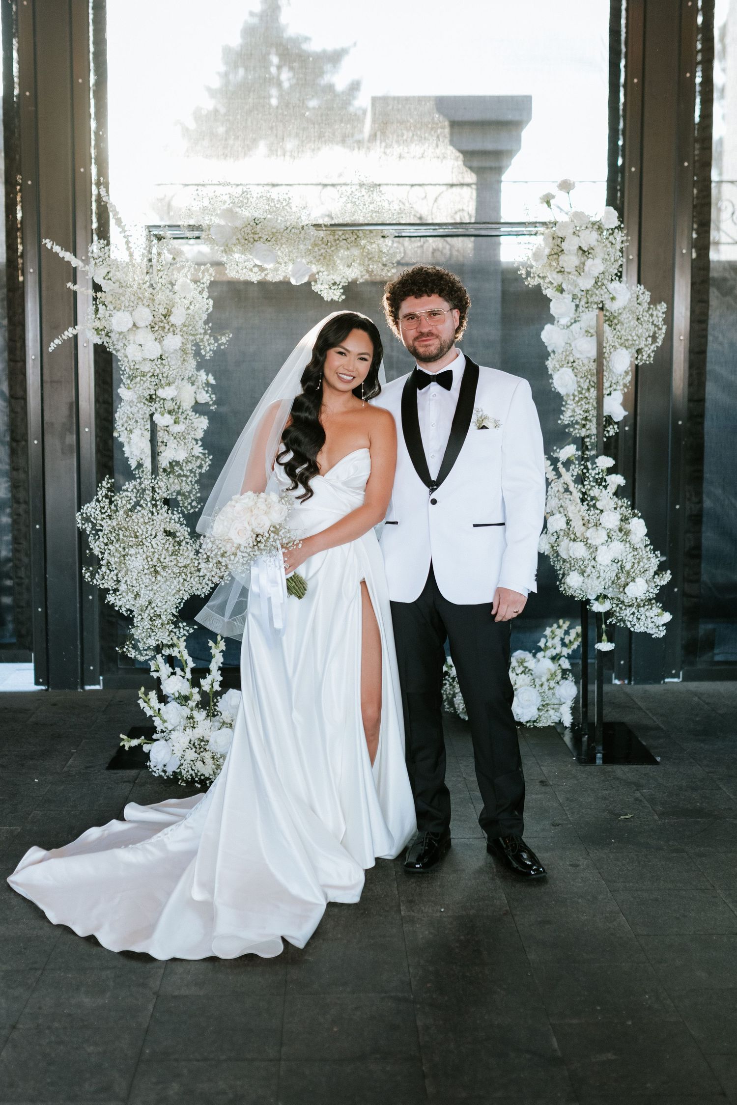 Wedding portrait in front of white floral arch with modern architecture in background.