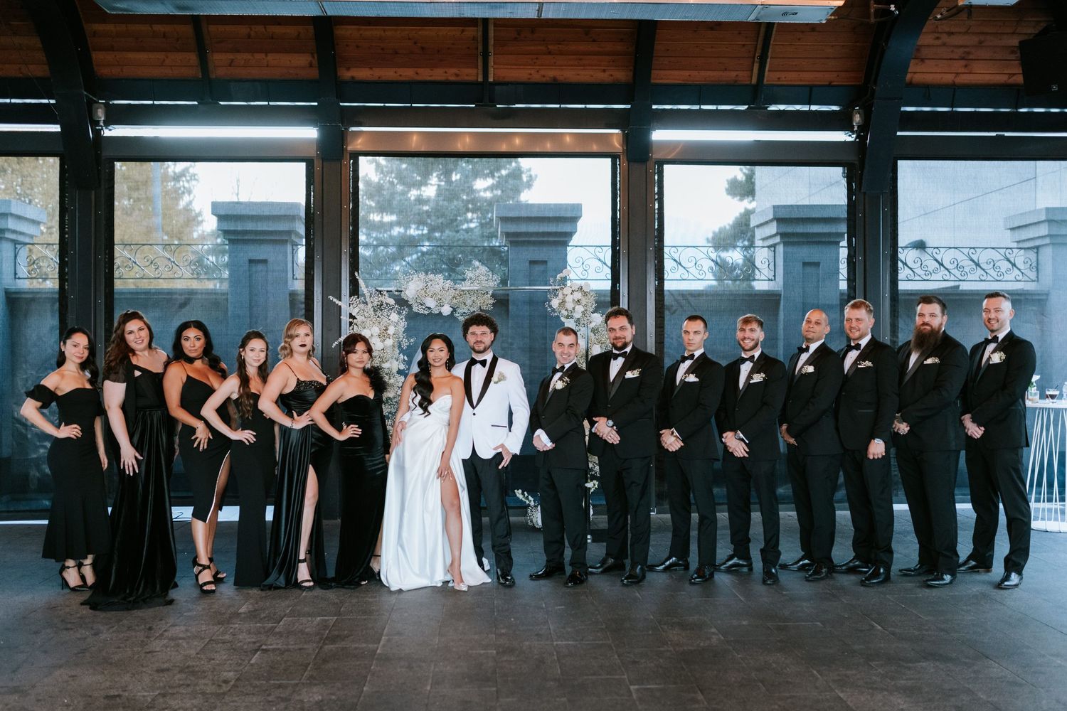 Wedding party lined up in elegant black attire with white dress in center against modern industrial backdrop.