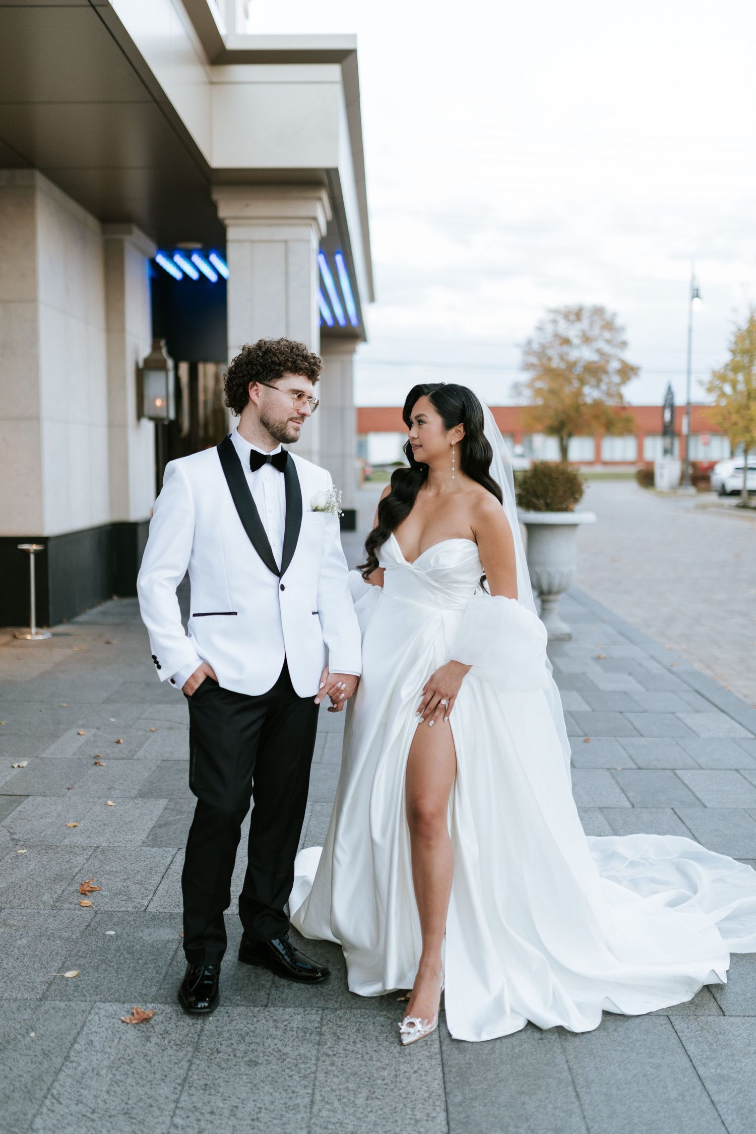 Elegant couple in white tuxedo and flowing wedding gown pose on city sidewalk.