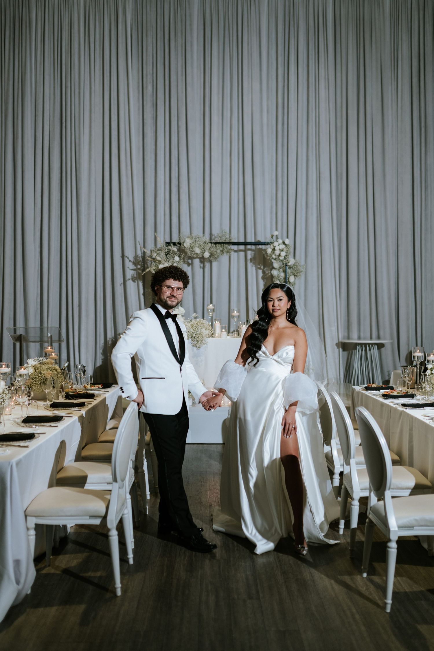 Newlyweds pose together in an elegant white wedding reception setup with floral arch and draped backdrop.