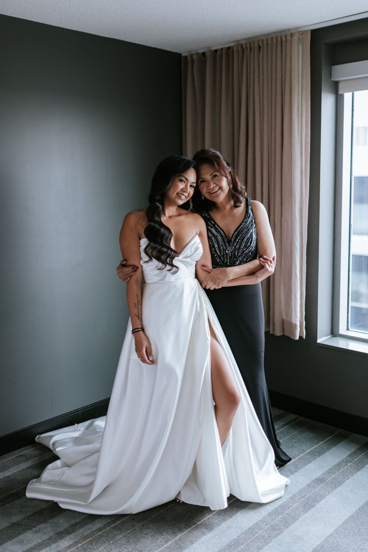 Two people embrace while wearing formal dresses in a room with dark green walls and beige curtains.
