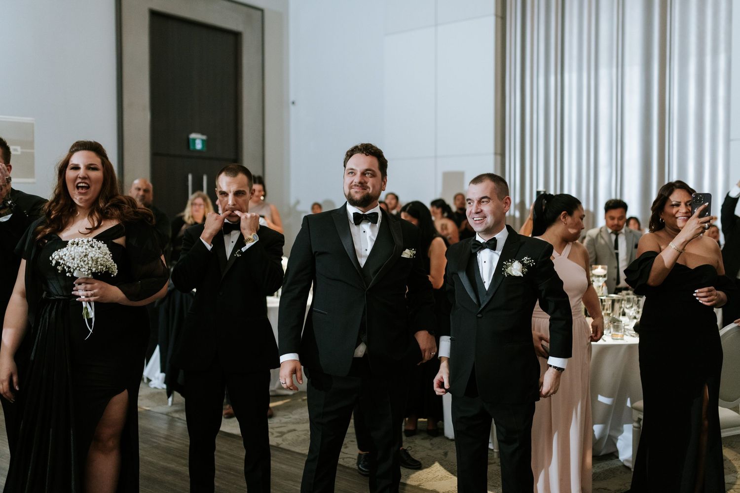 Wedding party in black formal attire walking and smiling during reception entrance.
