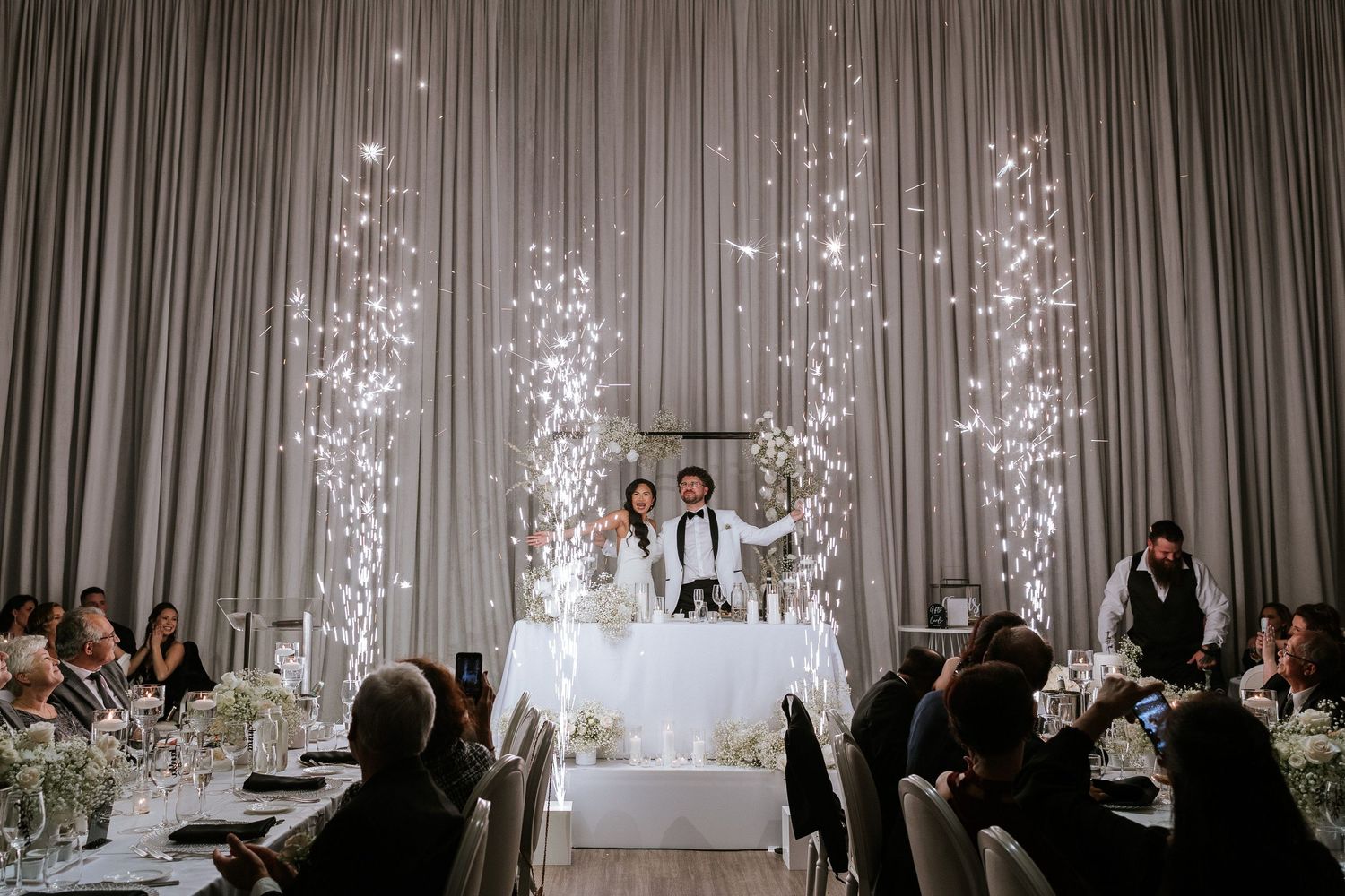 Wedding reception setup with dramatic sparkler effects and elegant grey draping behind head table.