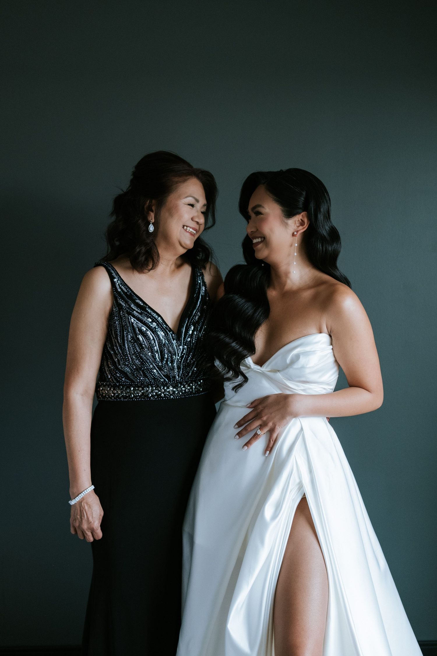 Two people in elegant evening wear share a candid moment against a dark wall.