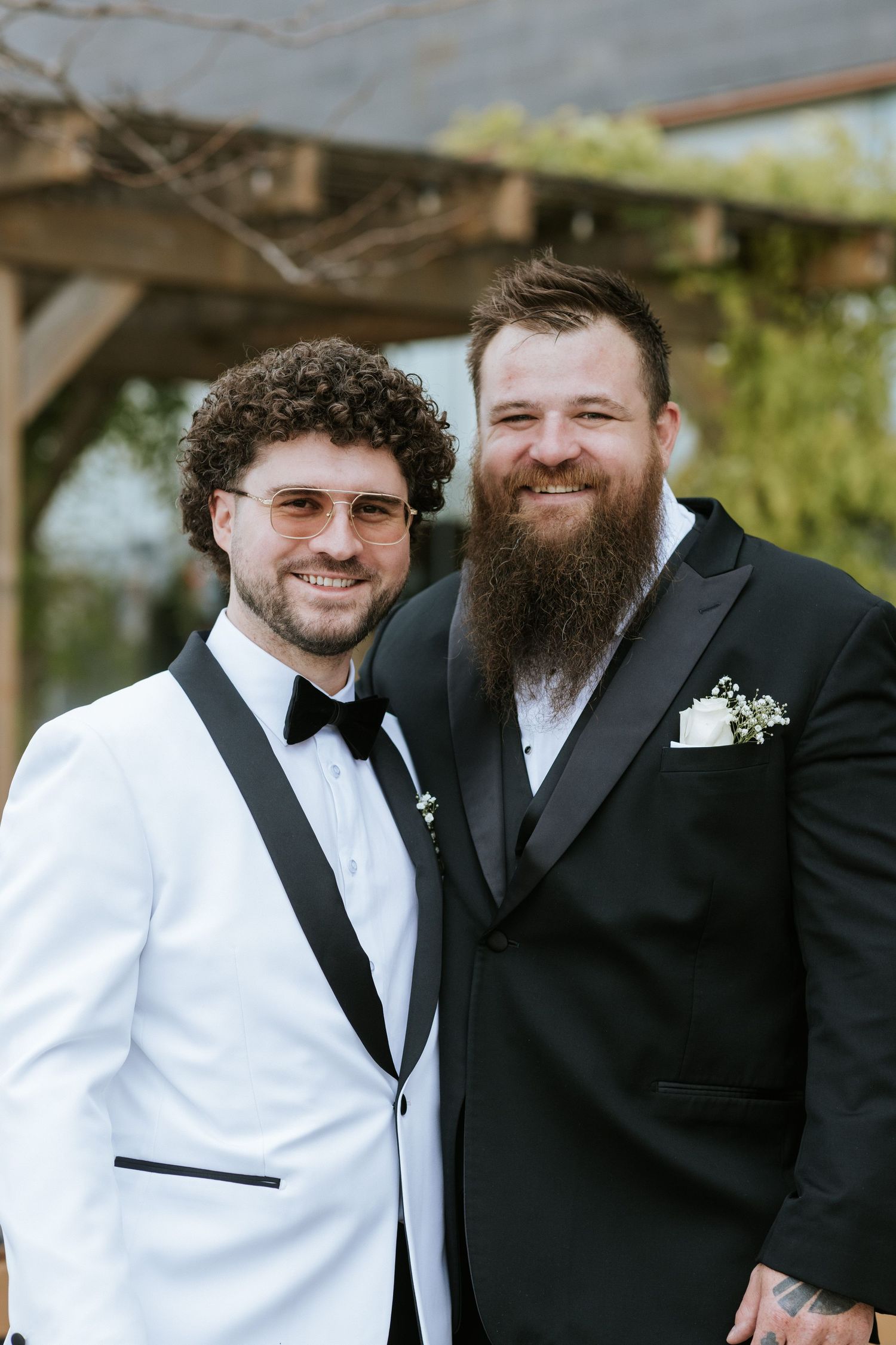 Two people in formal attire pose together under a wooden pergola.