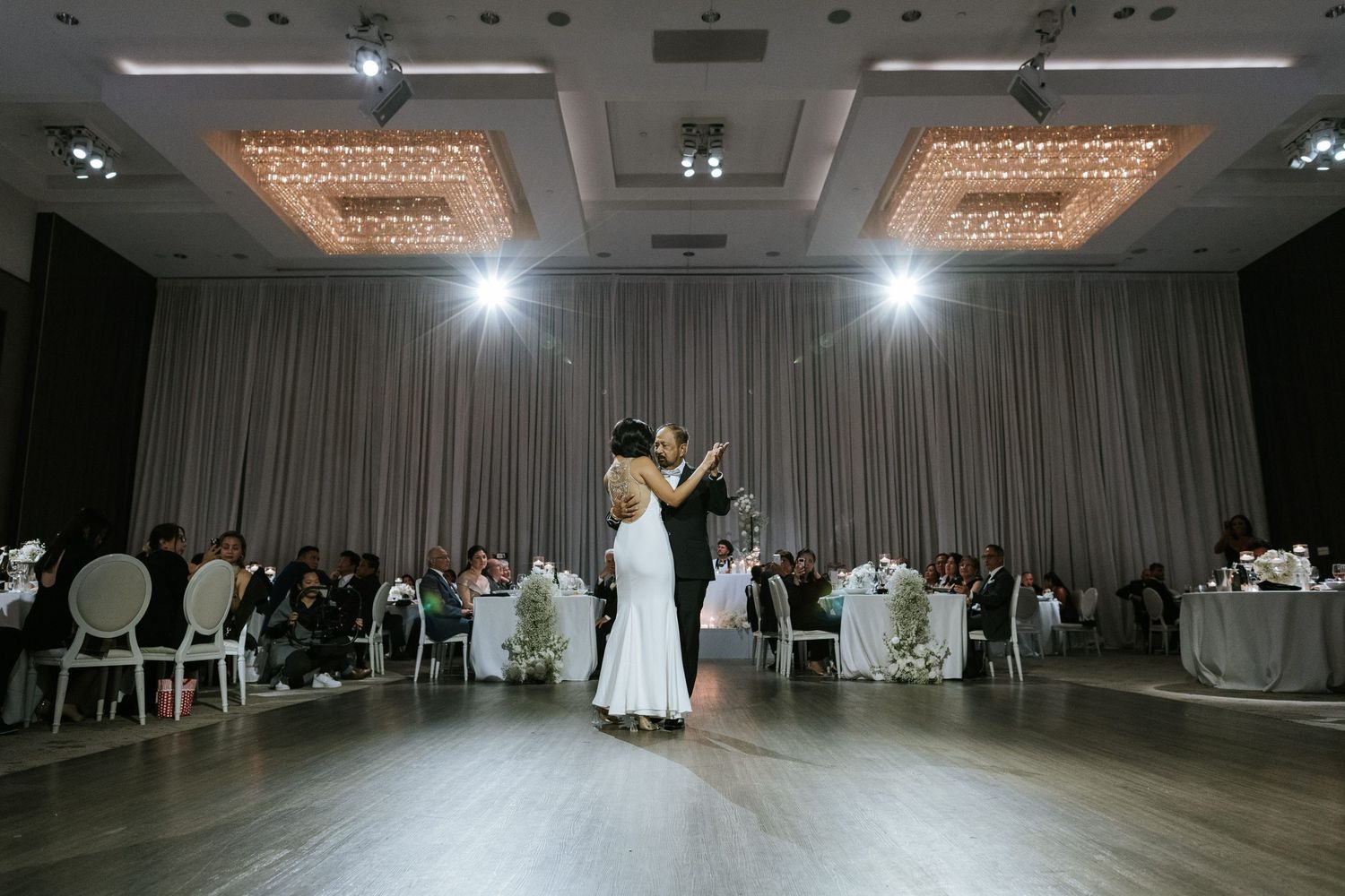 Couple sharing first dance in elegant ballroom with dramatic lighting and reception setup.