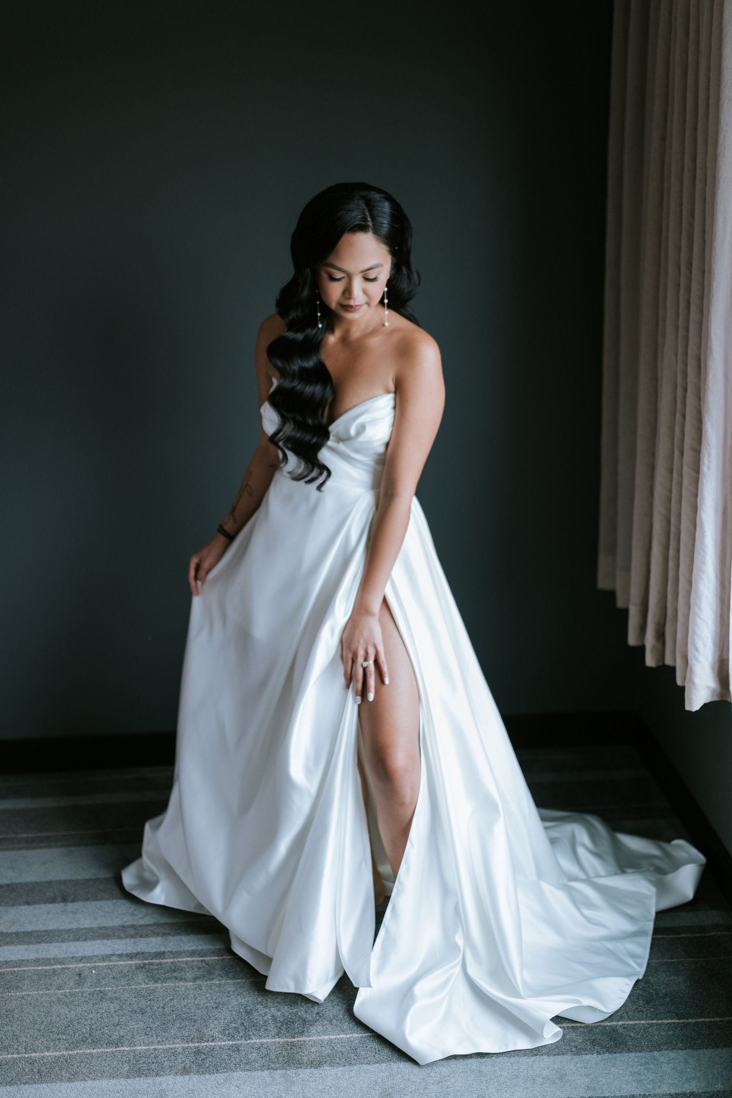 A person in a flowing white wedding gown with thigh-high slit poses against a dark wall.
