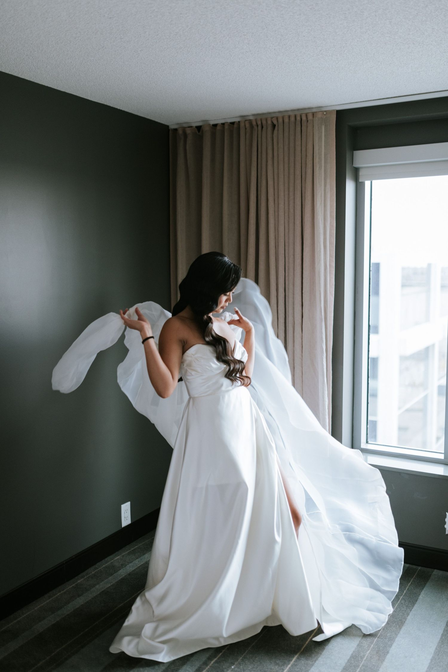 A bride in a flowing white wedding dress dances elegantly in a room with dark walls and natural window light.