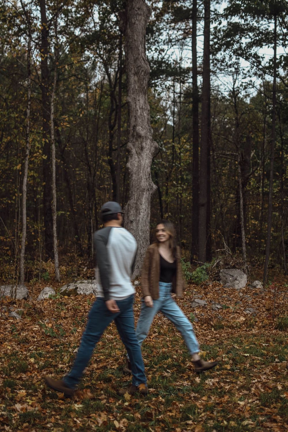 Two people walking through autumn woods with fallen leaves on forest floor, captured in moody lighting.
