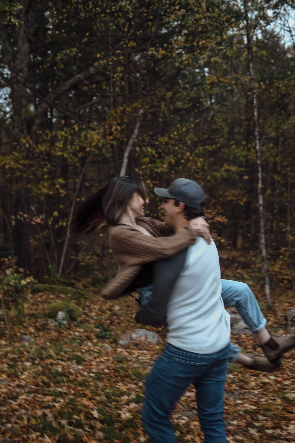 A couple embraces playfully in autumn woods with fallen leaves on the ground and trees in the background.