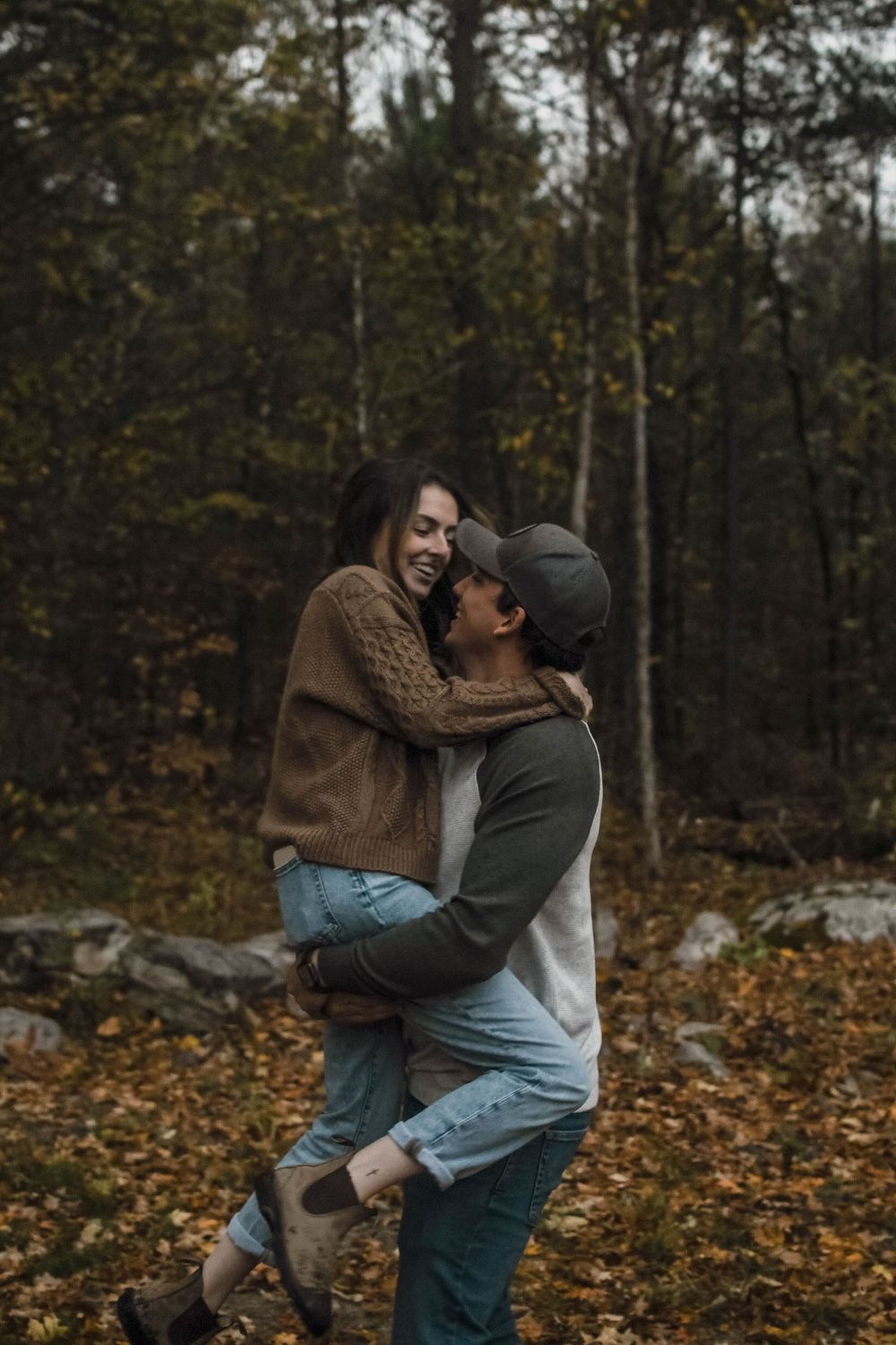 A couple shares an intimate moment embracing and laughing together in a forest during autumn with fallen leaves.