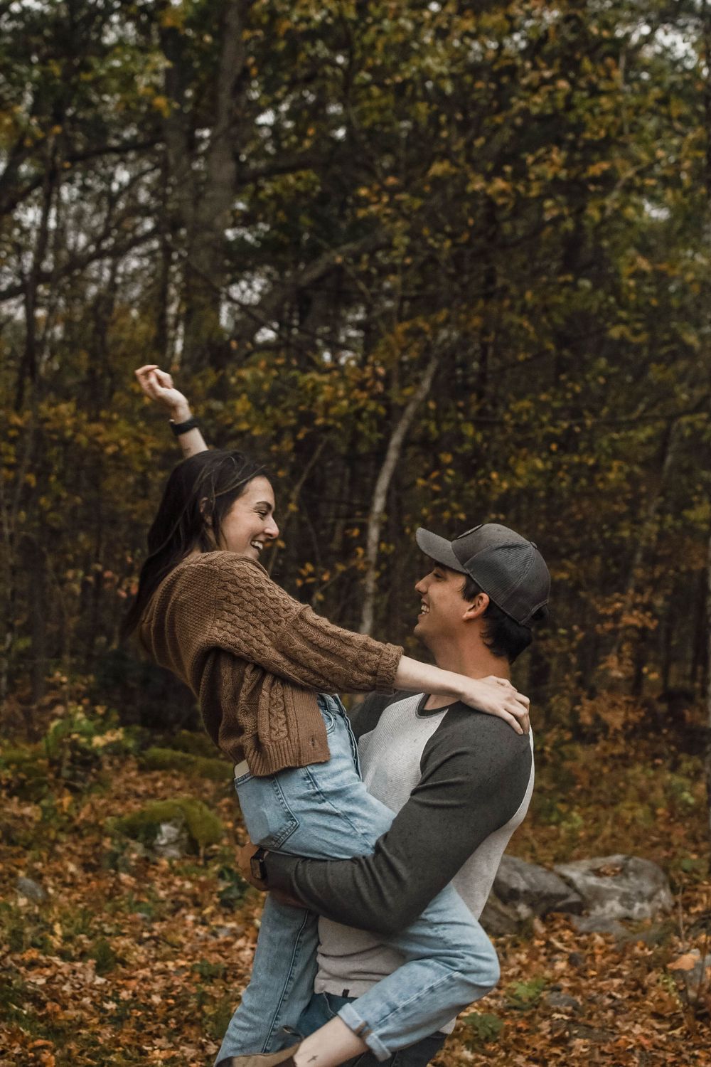 A couple embraces joyfully in an autumn forest setting with fallen leaves and trees in the background.