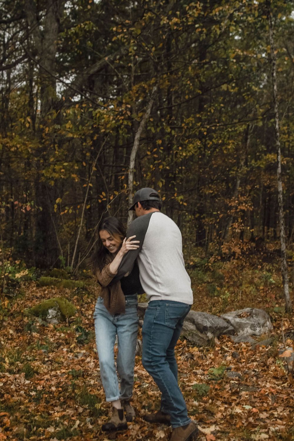 A couple playfully interacts amidst fallen autumn leaves in a moody forest setting.