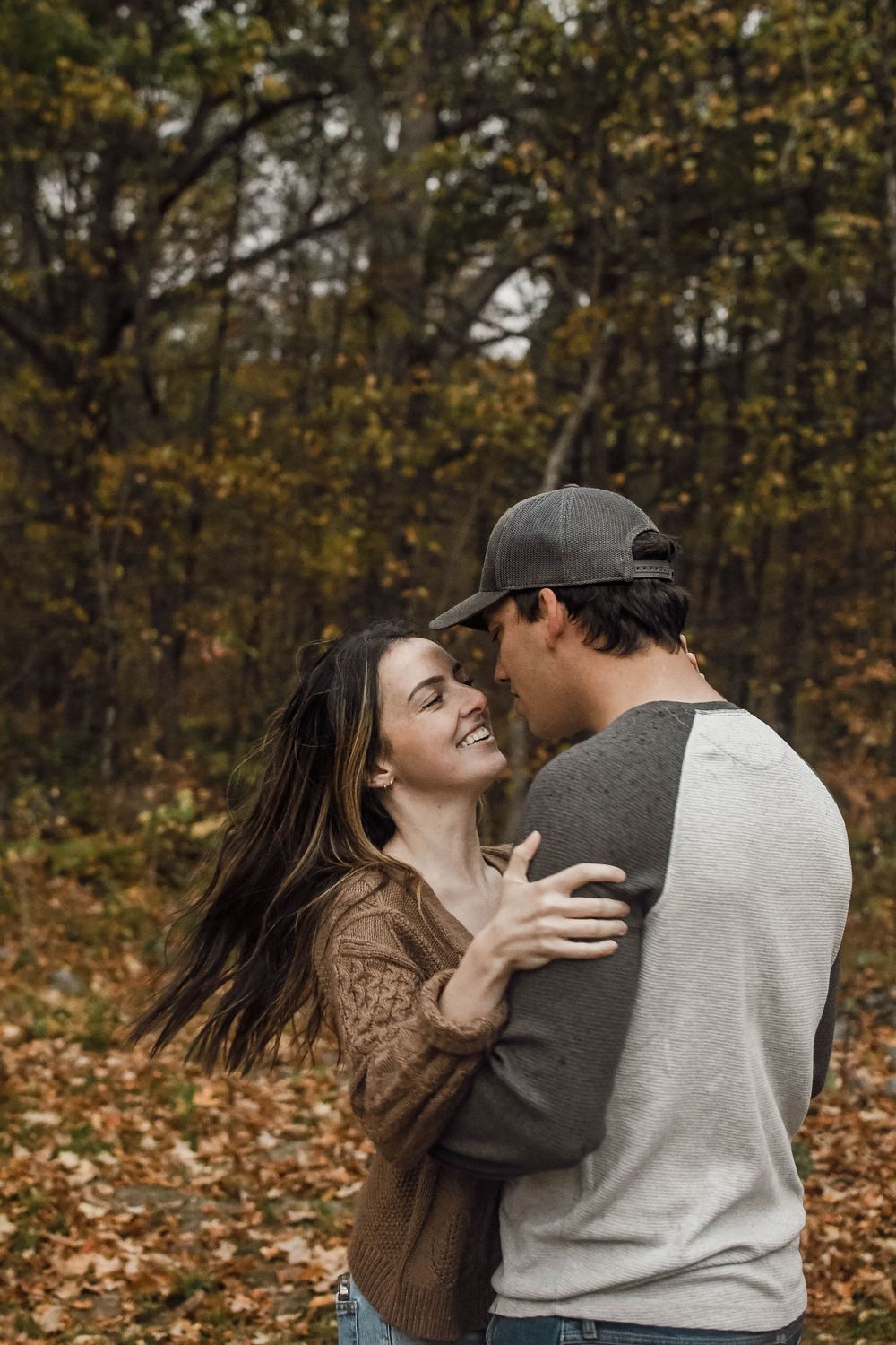 A couple embraces and laughs together in an autumn forest setting with fallen leaves on the ground.