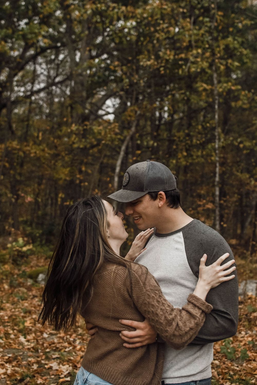 A couple embraces in an autumn forest setting surrounded by fallen leaves and trees.