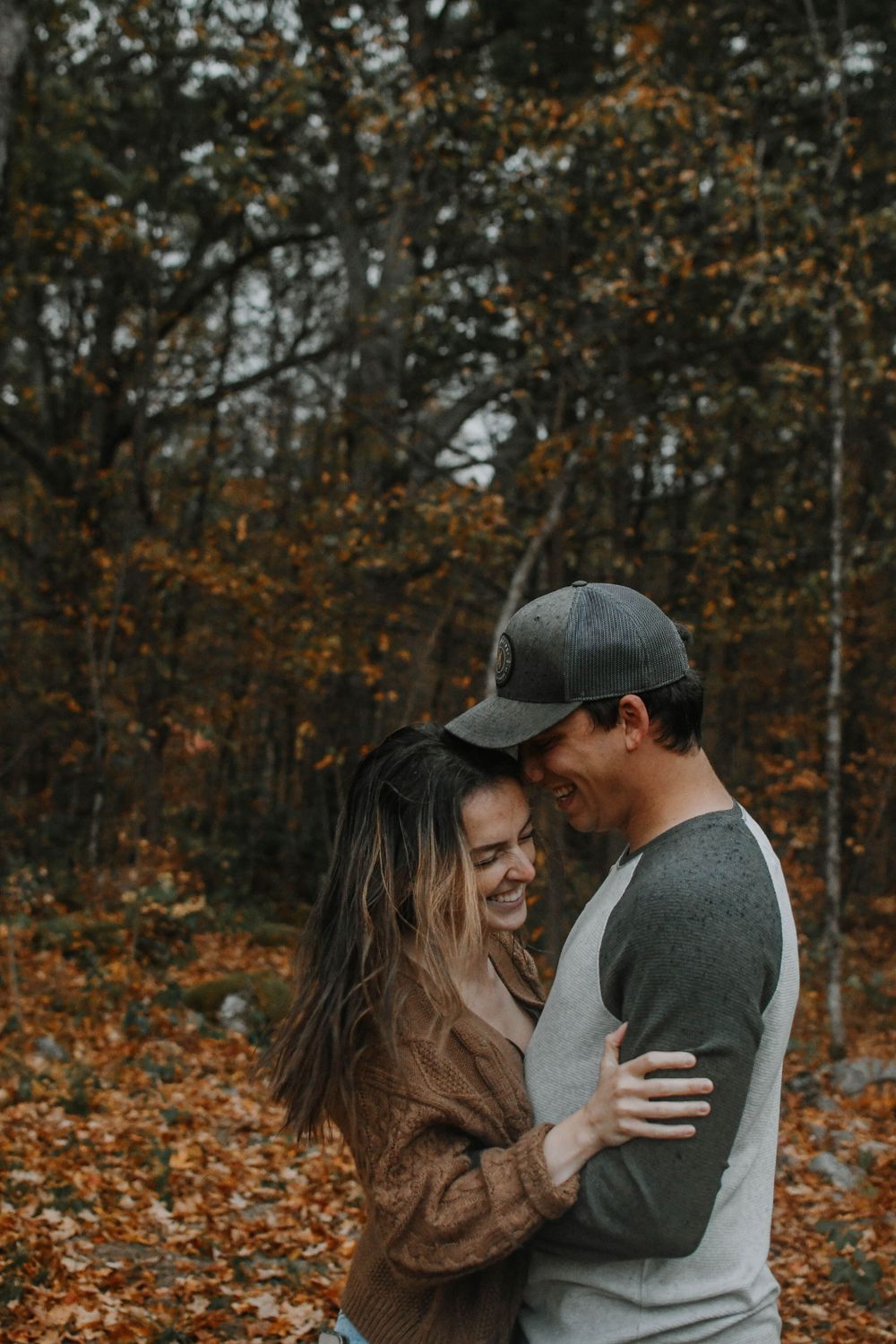 A couple embraces and laughs together in an autumn forest with orange fallen leaves on the ground.