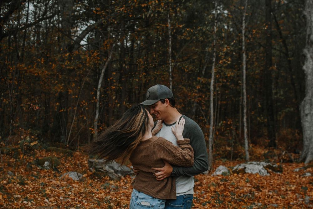 Couple embraces in romantic autumn forest setting surrounded by fallen orange leaves and bare trees.