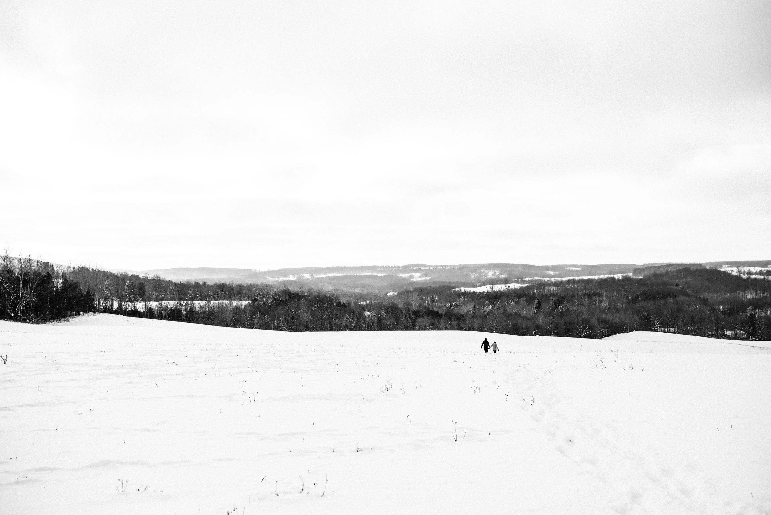 Two distant figures traverse a snowy winter hillside overlooking a vast rural landscape in black and white.