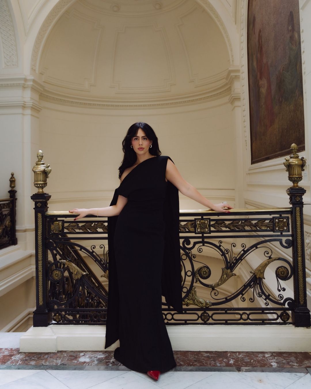 Elegant woman in a black one-shoulder gown posing on an ornate staircase in a grand venue.