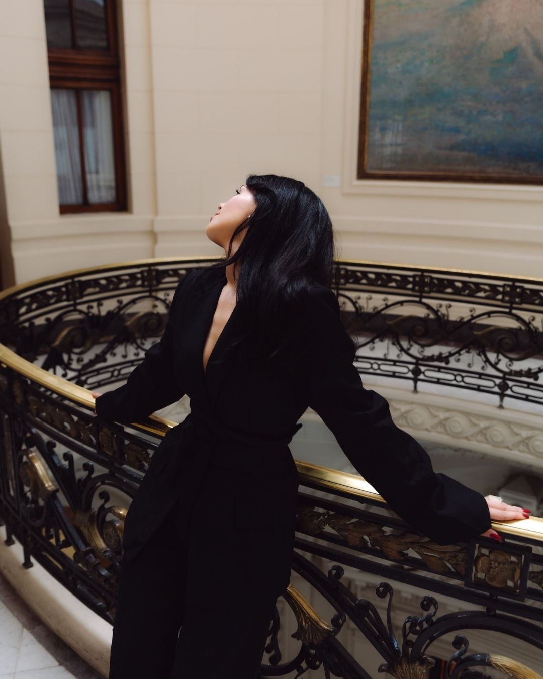 Woman in black deep-back blazer leaning on ornate gold staircase railing in elegant interior.
