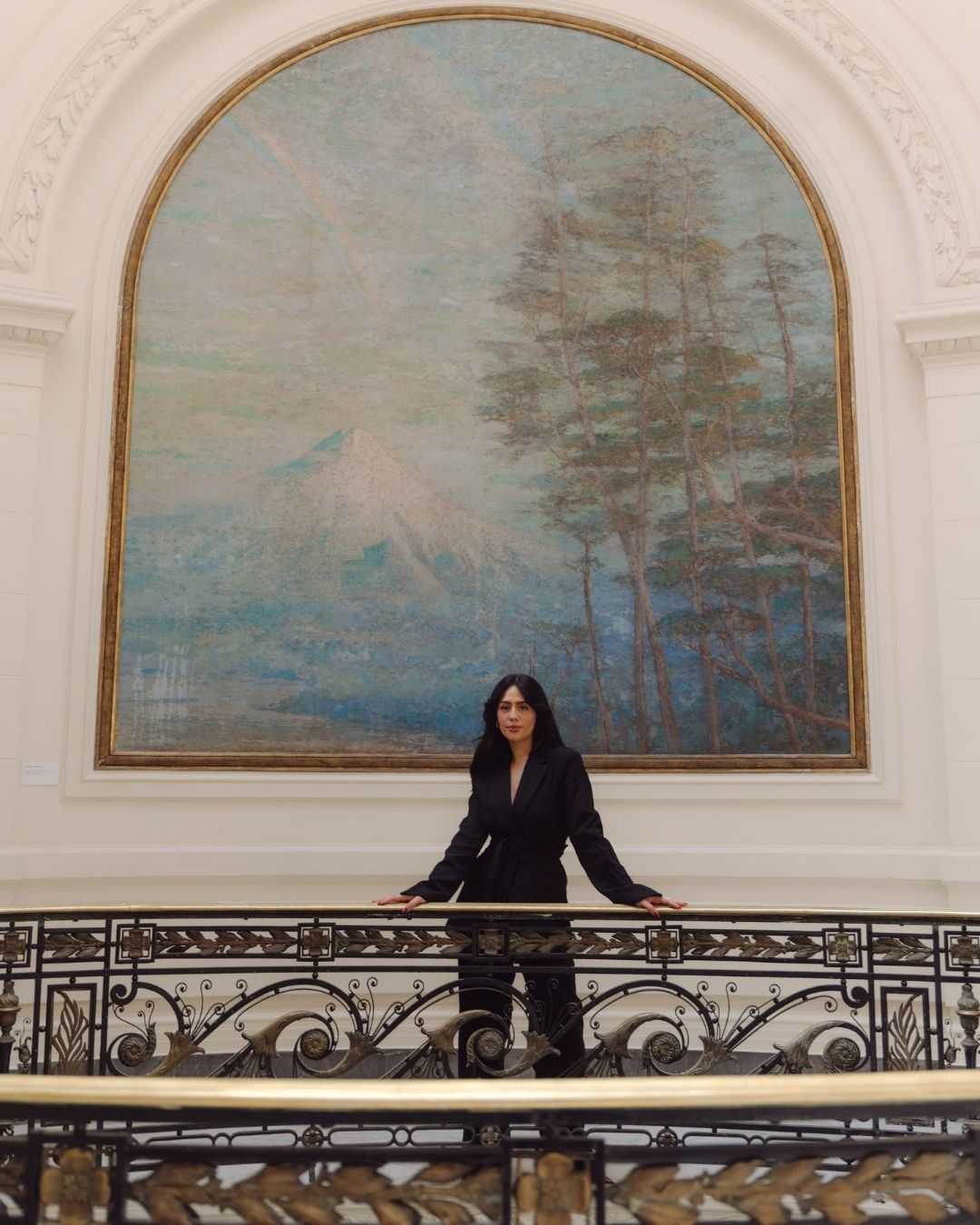 Woman in black suit standing on balcony before large arched mountain landscape oil painting.