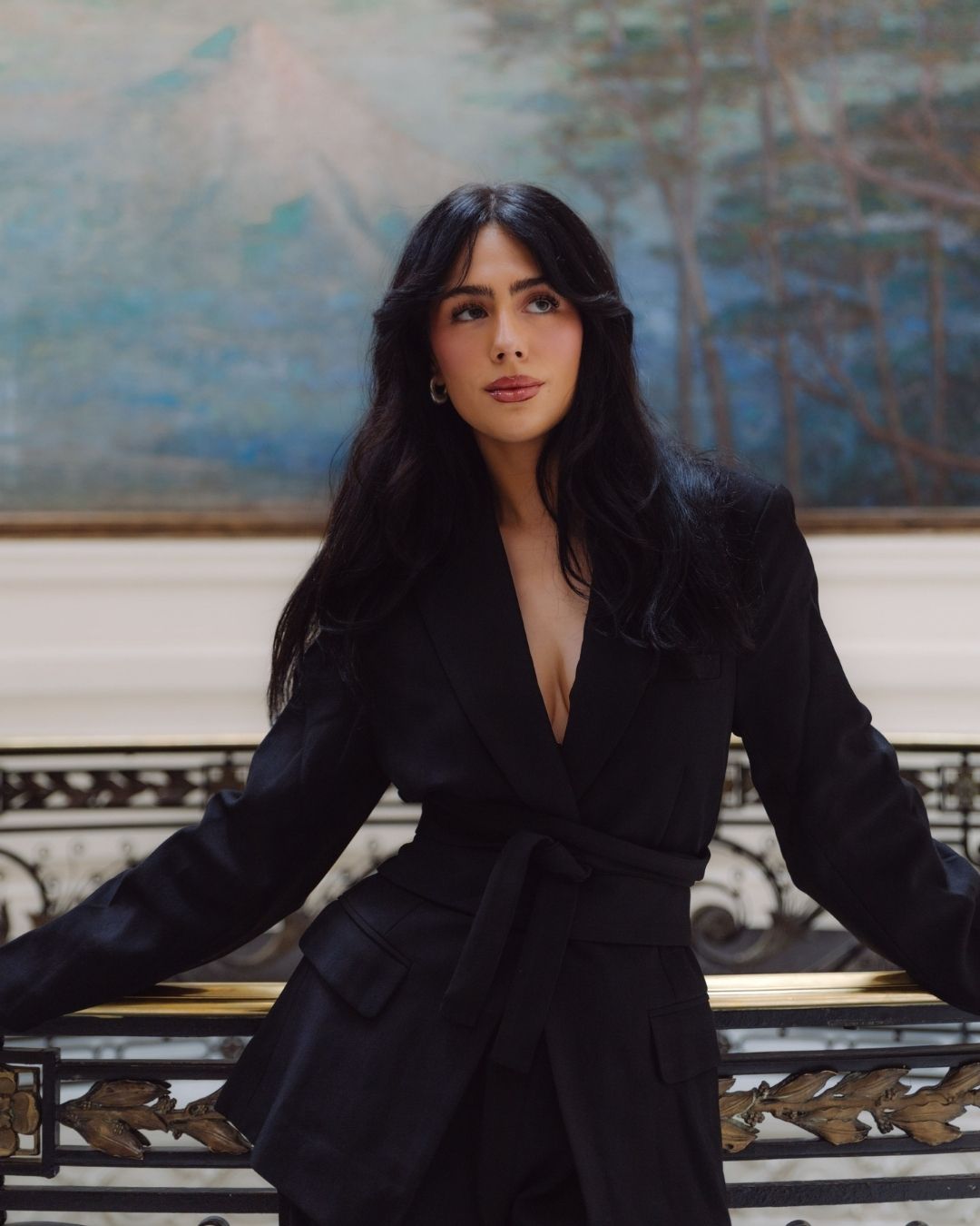 Woman in black wrap blazer posing confidently on ornate gold railing staircase balcony.