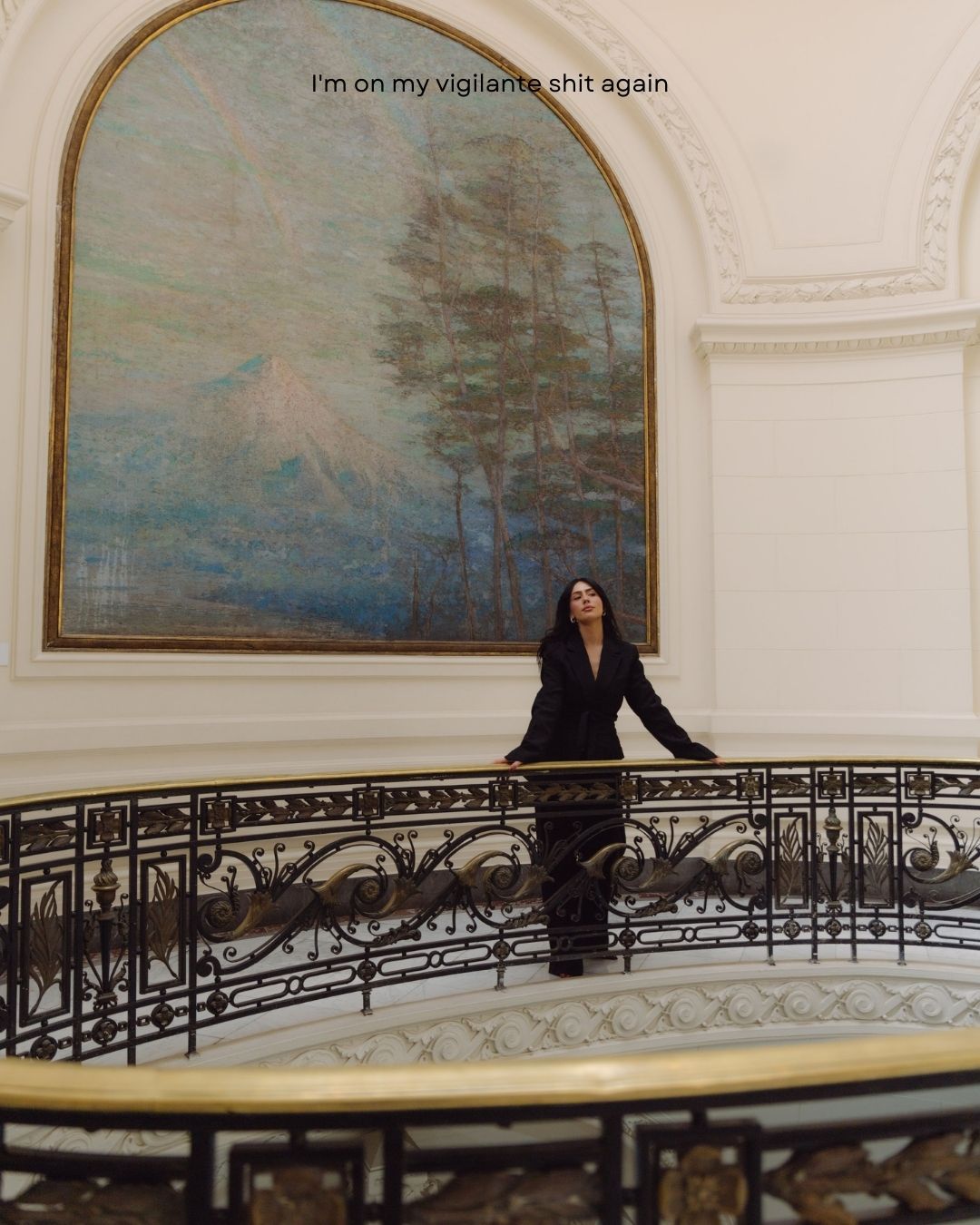 Woman in black suit gazing upward on ornate balcony with large arched landscape painting.