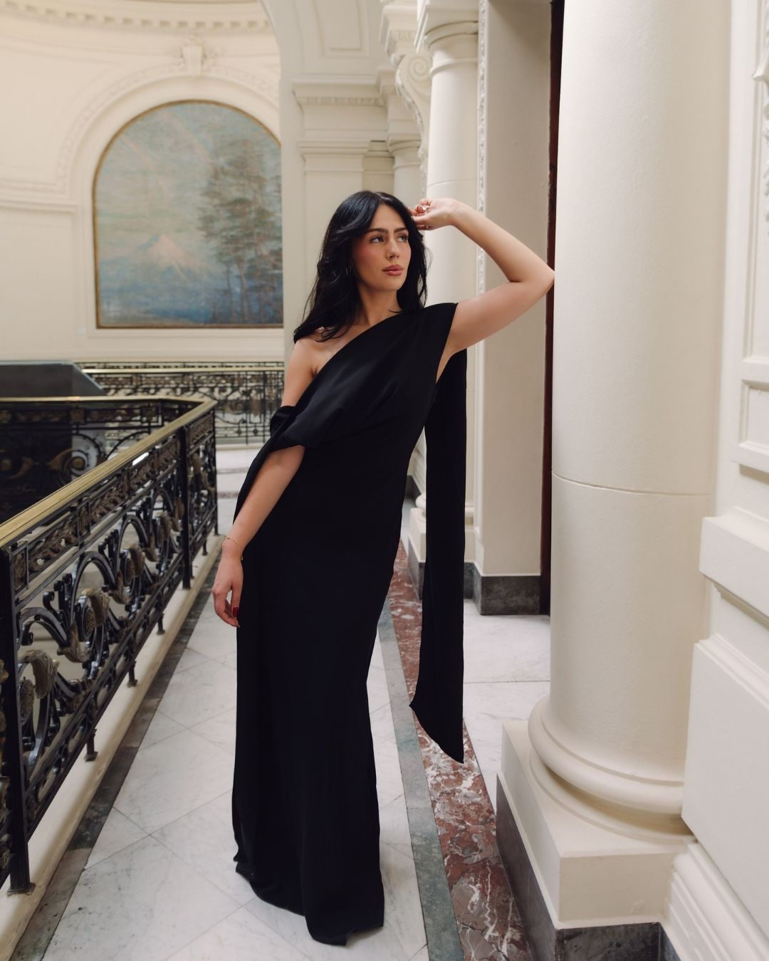 Woman in an elegant black one-shoulder floor-length gown posing in a grand classical hallway.