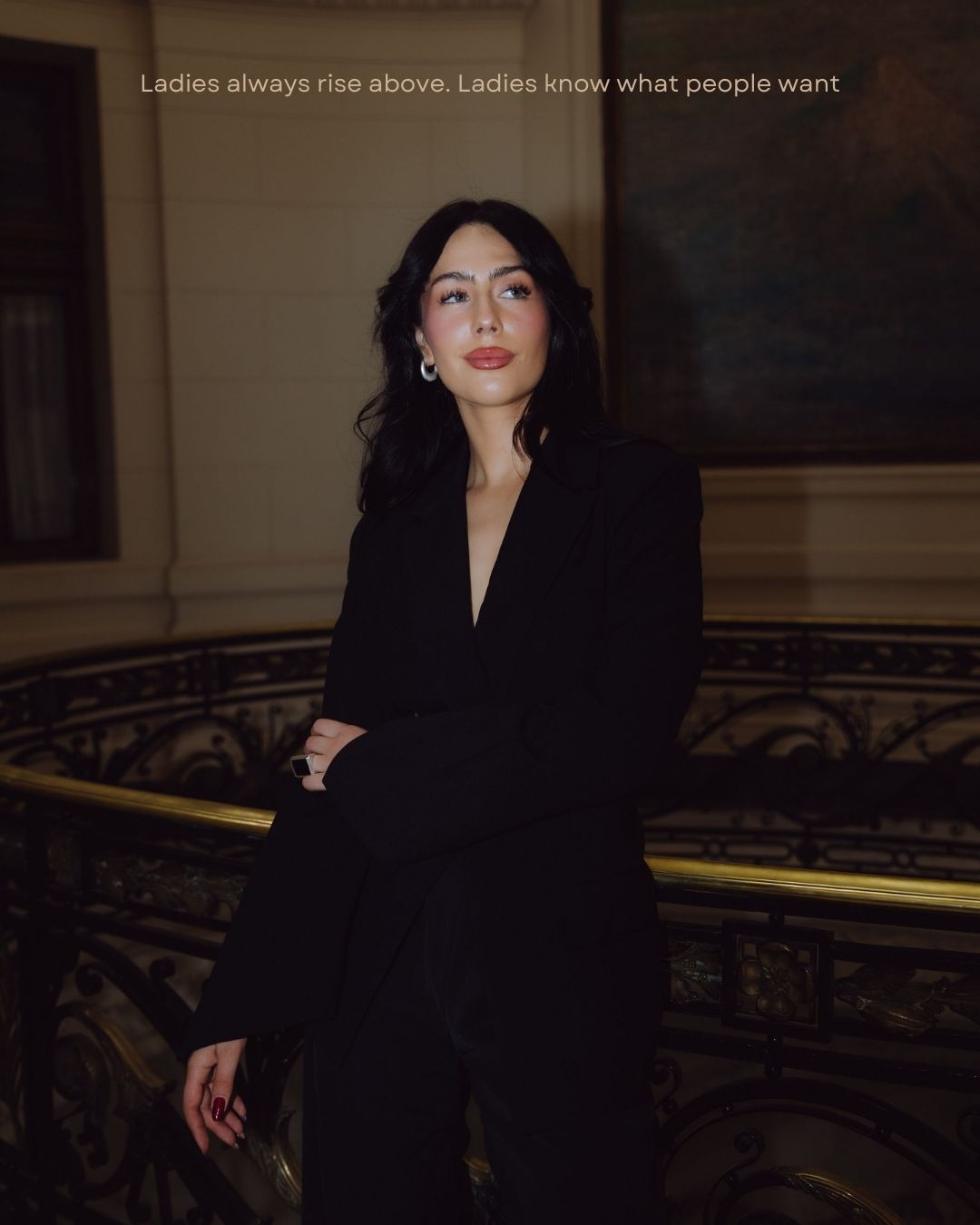 Woman in black blazer suit posing on luxury staircase with gold railings and oil painting.