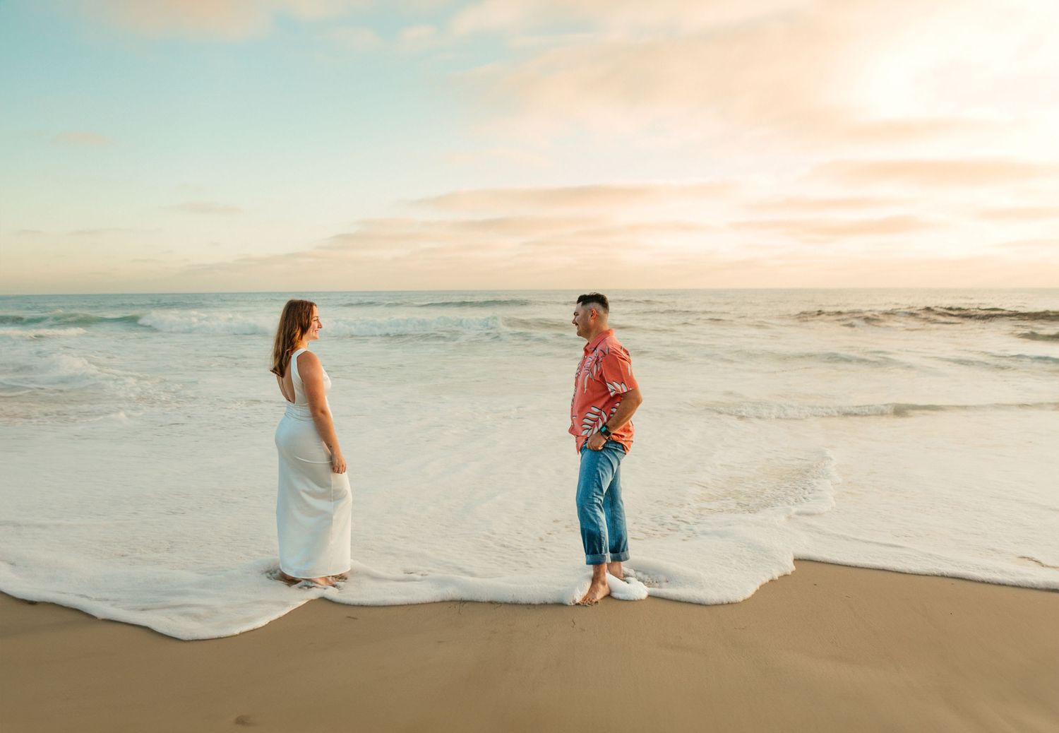 Couple stands facing each other on sandy beach during golden hour.