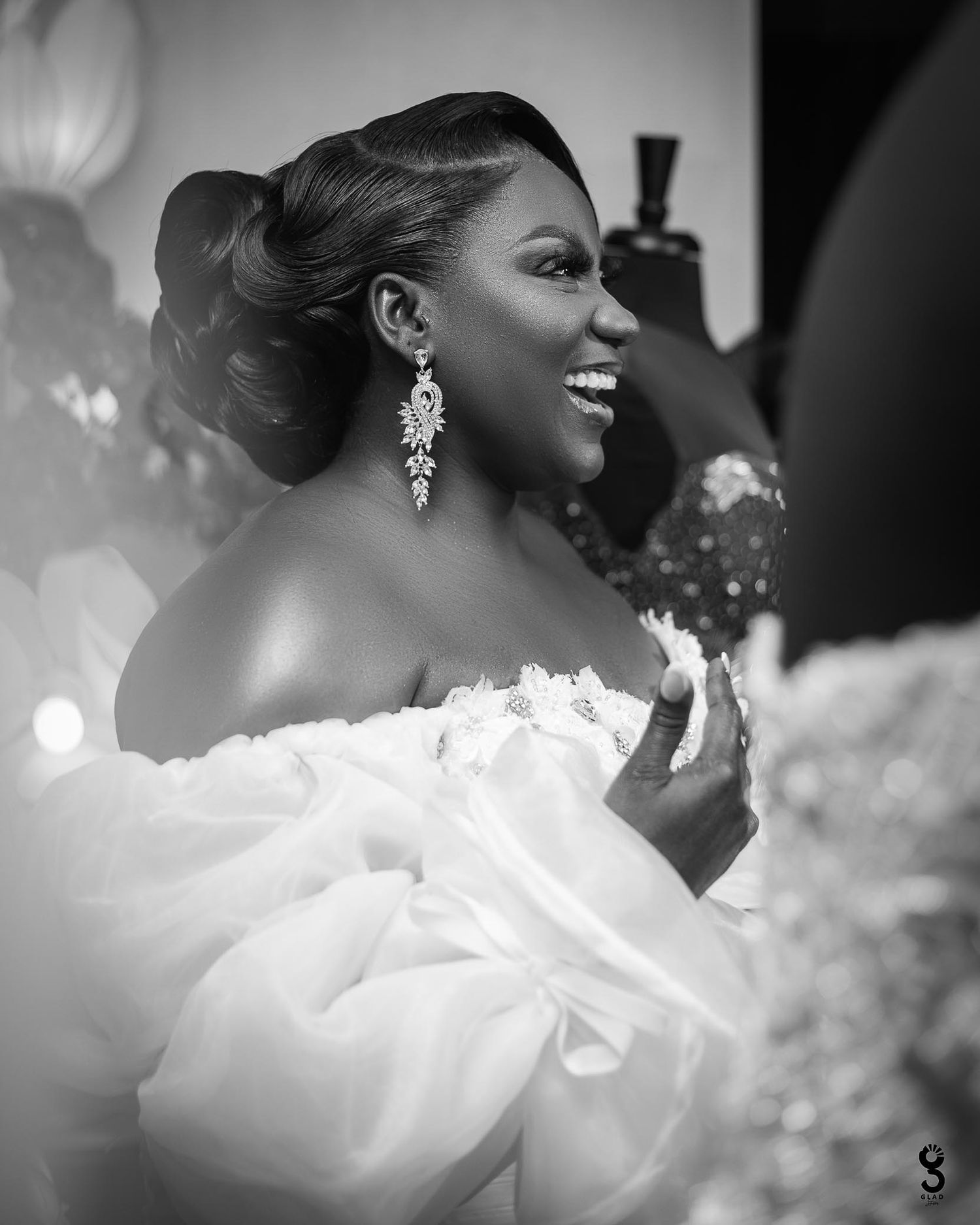 A radiant bride in an off-shoulder white gown holds a beautiful bouquet of white flowers while standing by a window.