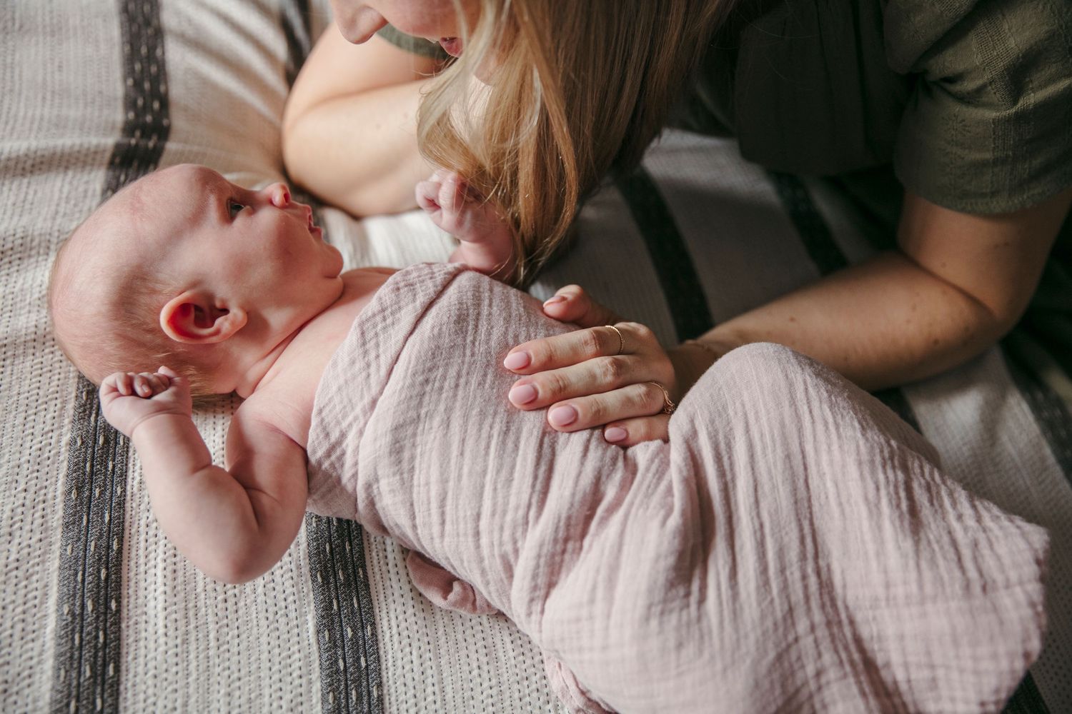 Baby wrapped in soft muslin fabric lying on a striped blanket with gentle care. Mom leans over baby with hand on baby's belly.
