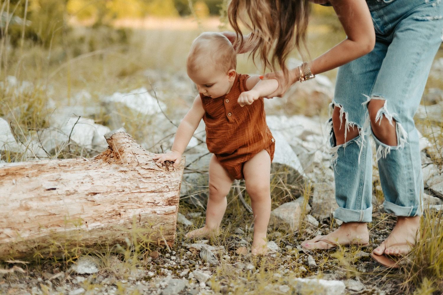 Child in brown romper explores rocky terrain while being supported on outdoor adventure at golden hour.