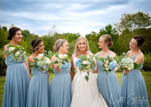 Elegant wedding photography of a bride with her bridesmaids in flowing blue dresses, posing with bouquets in a scenic outdoor setting.