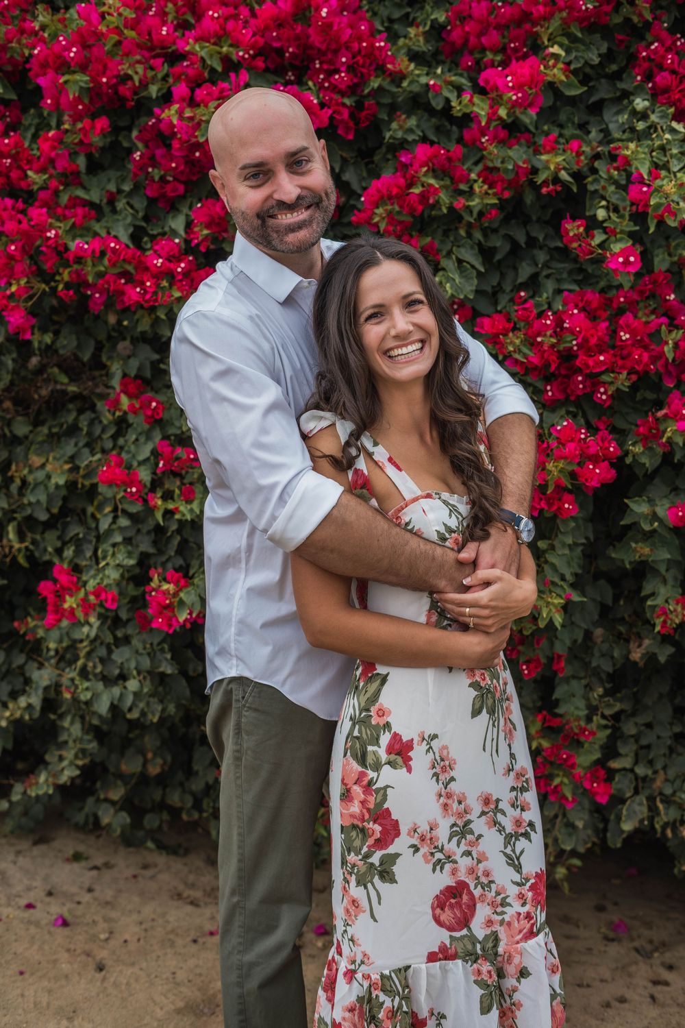 Couple embracing in front of vibrant pink bougainvillea bush during outdoor photoshoot.
