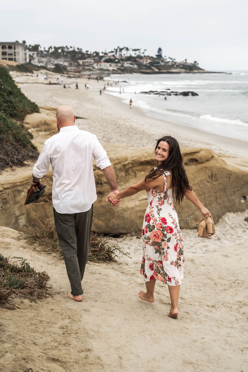 A couple strolling hand in hand on a scenic beach path with cliffs and ocean views in the background.