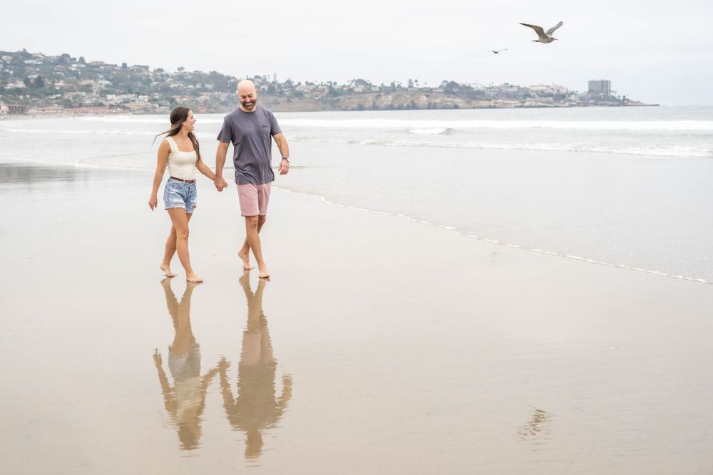 A couple walking barefoot on wet sand with their reflections visible and a seagull flying overhead.