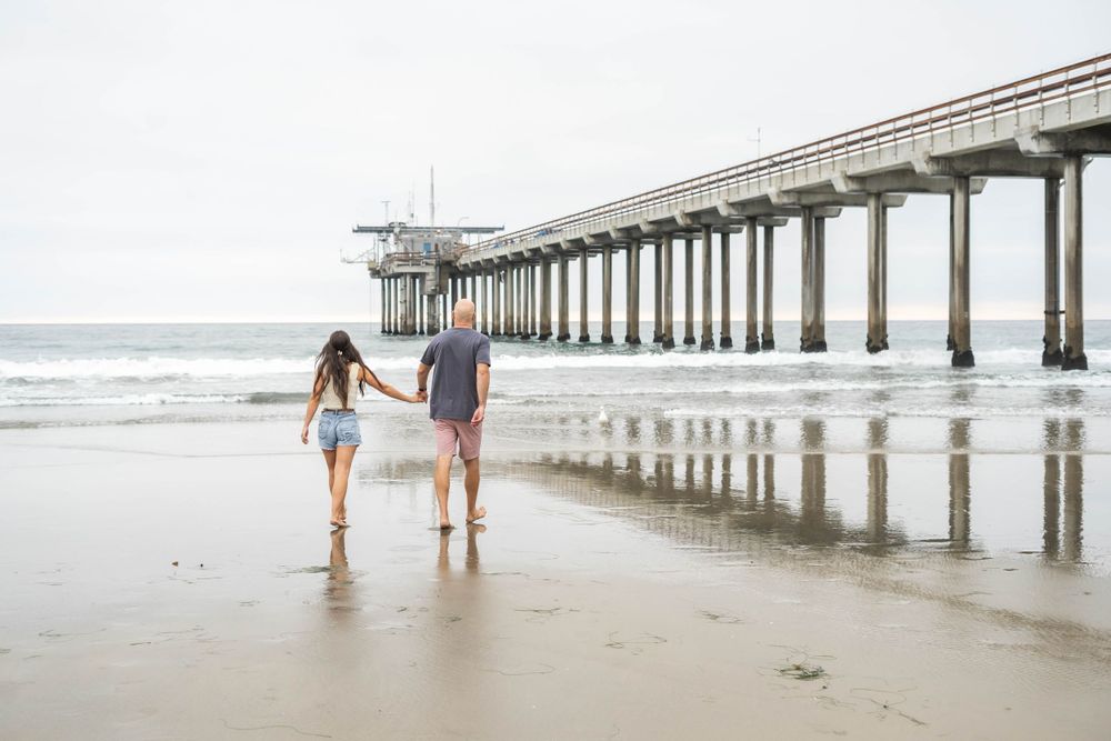 A couple walking barefoot along the shore near a long wooden pier.