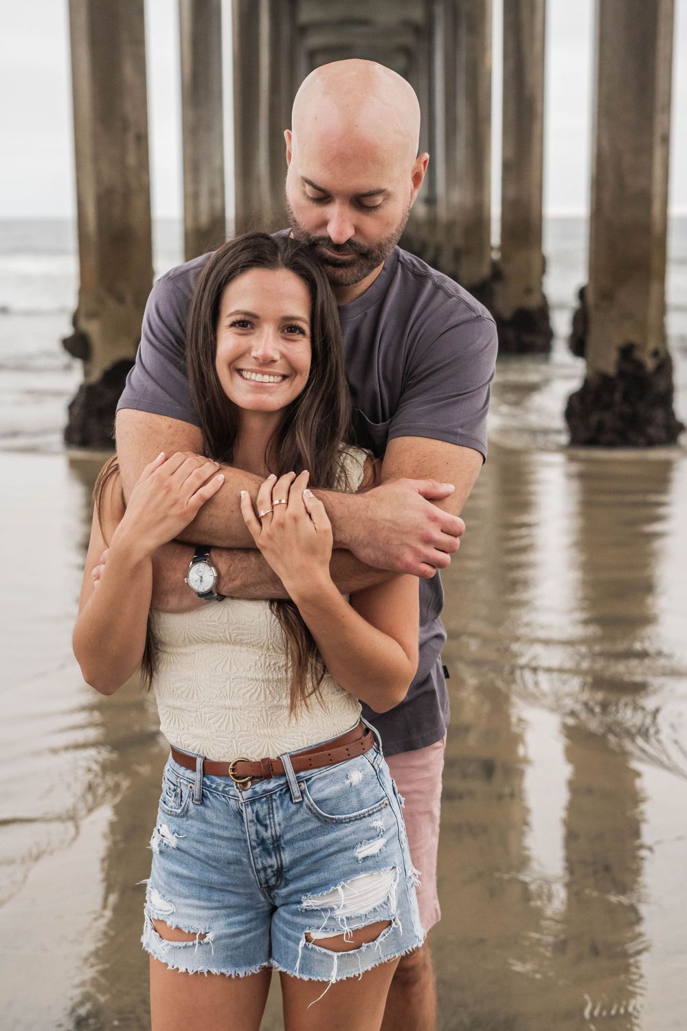 A romantic embrace under a beach pier with waves lapping at the shoreline.