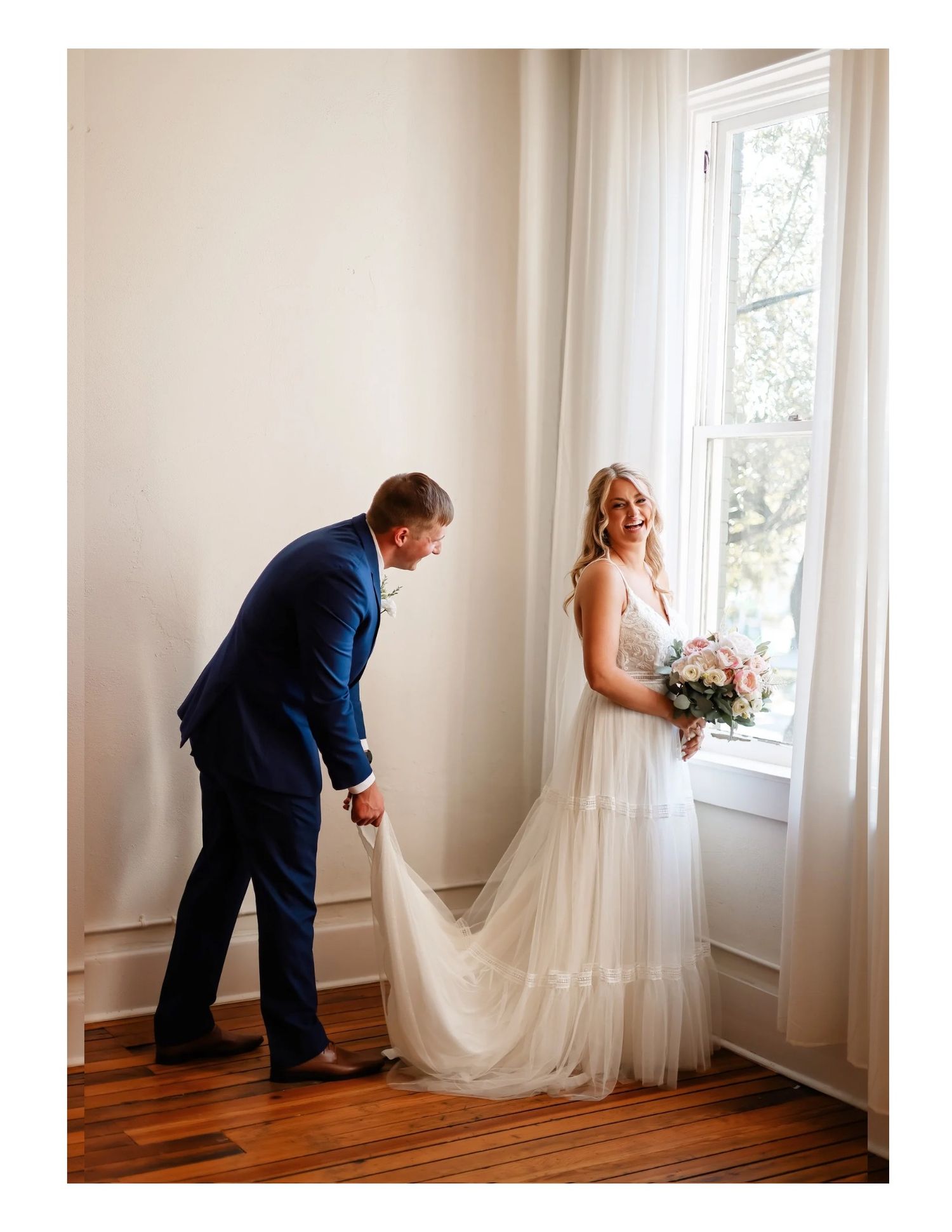 Bride in flowing white dress standing by window while dress train is adjusted.