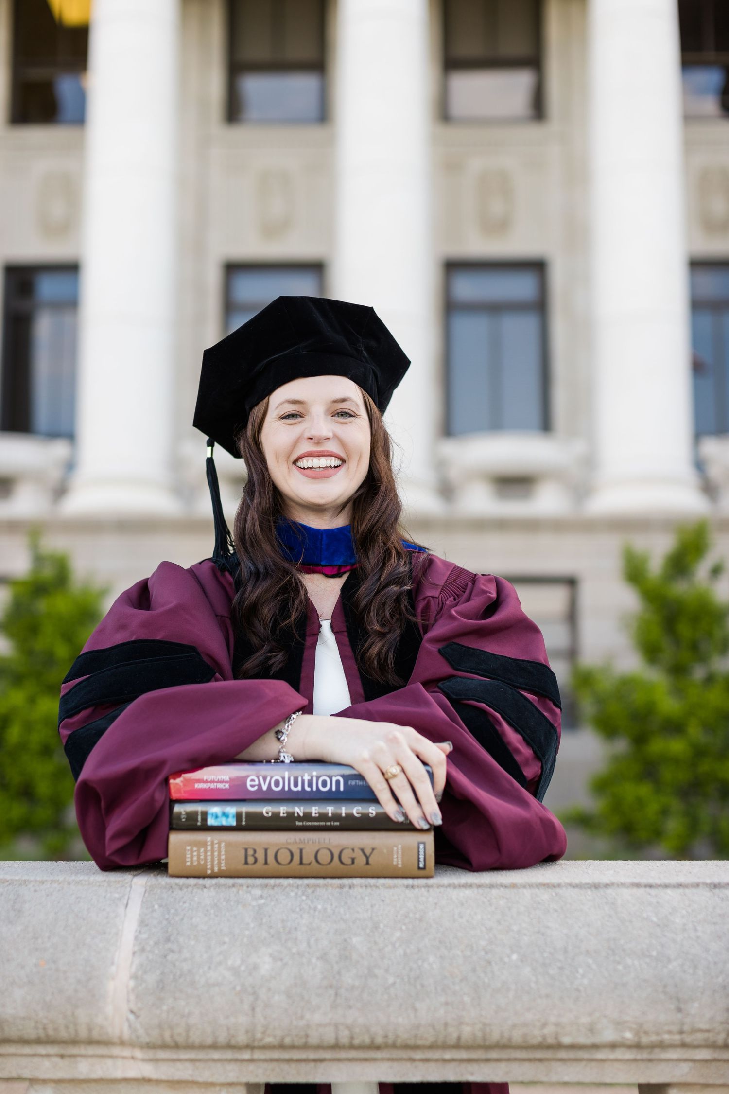 Graduate in burgundy academic regalia poses with textbooks including Evolution and Biology near classical columns.