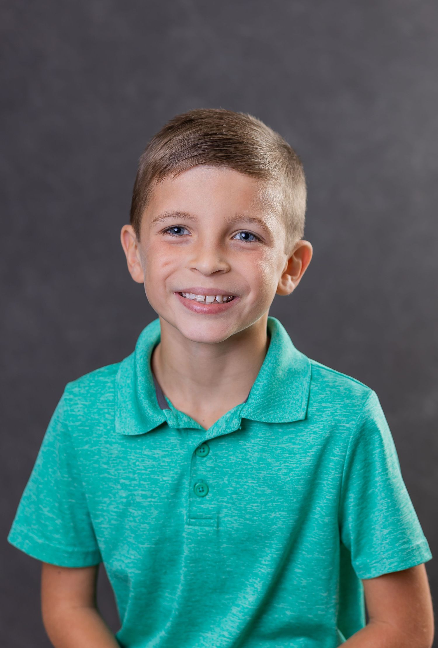 Young student in bright turquoise polo shirt sits for school portrait against gray background.