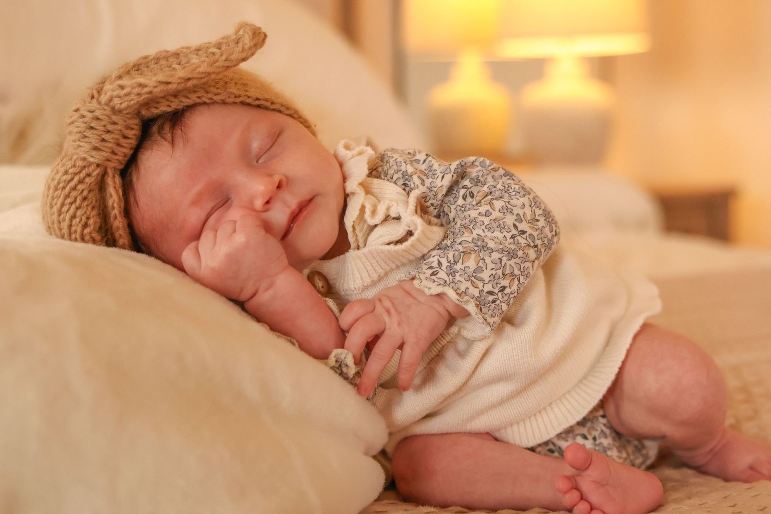 Newborn baby sleeping peacefully on cream-colored bedding while wearing a tan hat and floral print outfit.