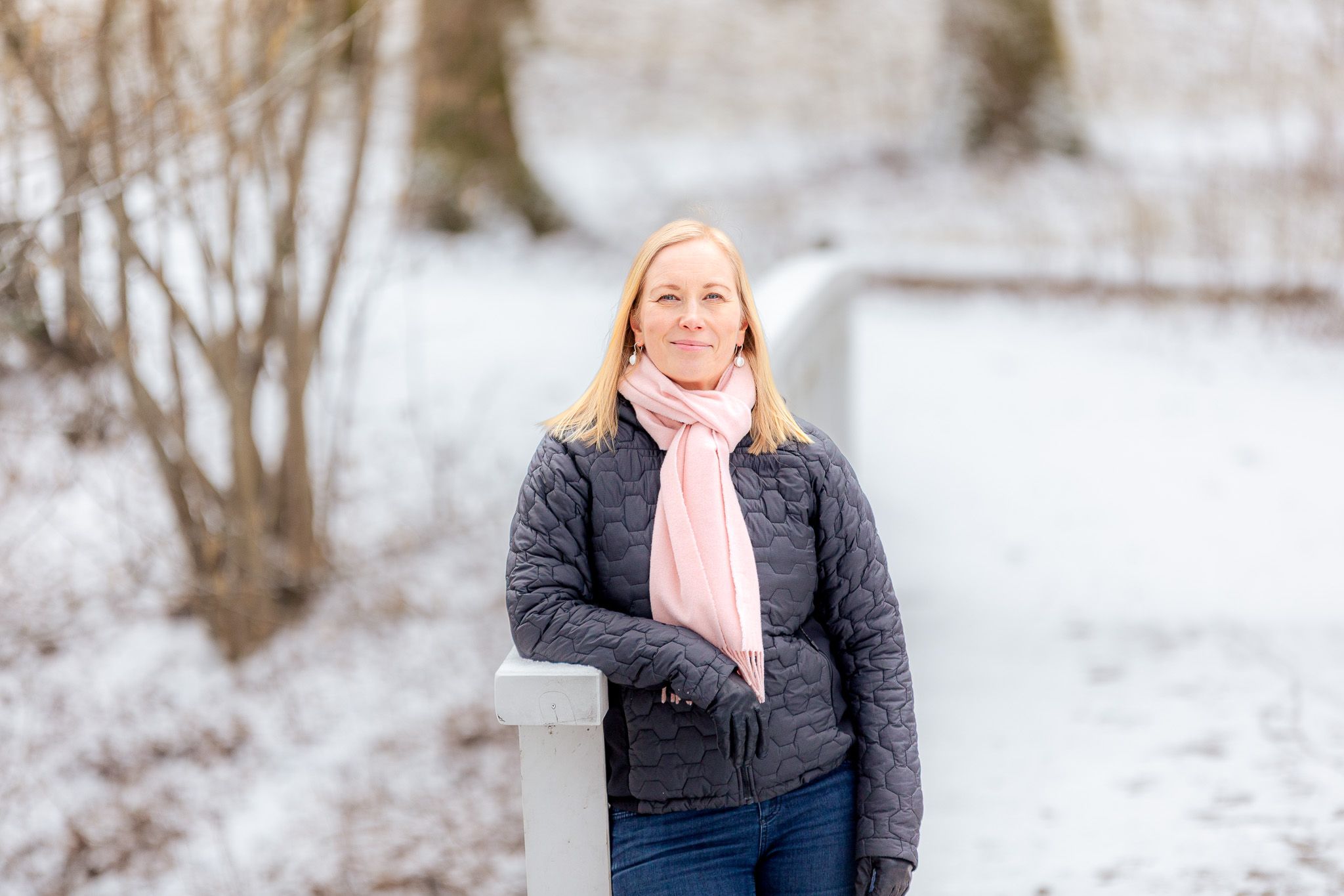 "Nainen seisoo lumisella sillalla käsi kaiteella, pukeutuneena tummaan takkiin, vaaleanpunaiseen huiviin ja farkkuihin."