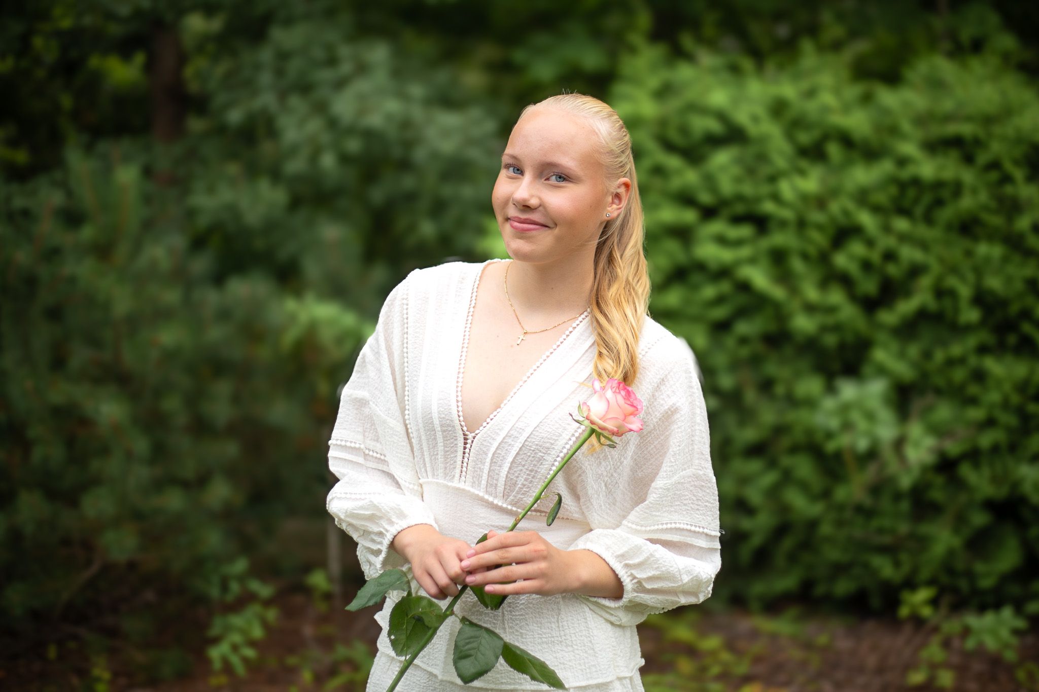 Nuori vaaleahiuksinen nainen seisoo vihreän pensaston edessä, pukeutuneena valkoiseen mekkoon ja pitelee vaaleanpunaista ruusua, hymyillen l