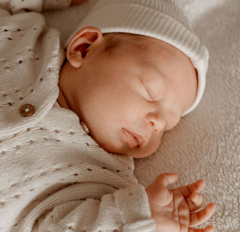Close-up profile view of a sleeping newborn wearing a white knit hat against a textured background.