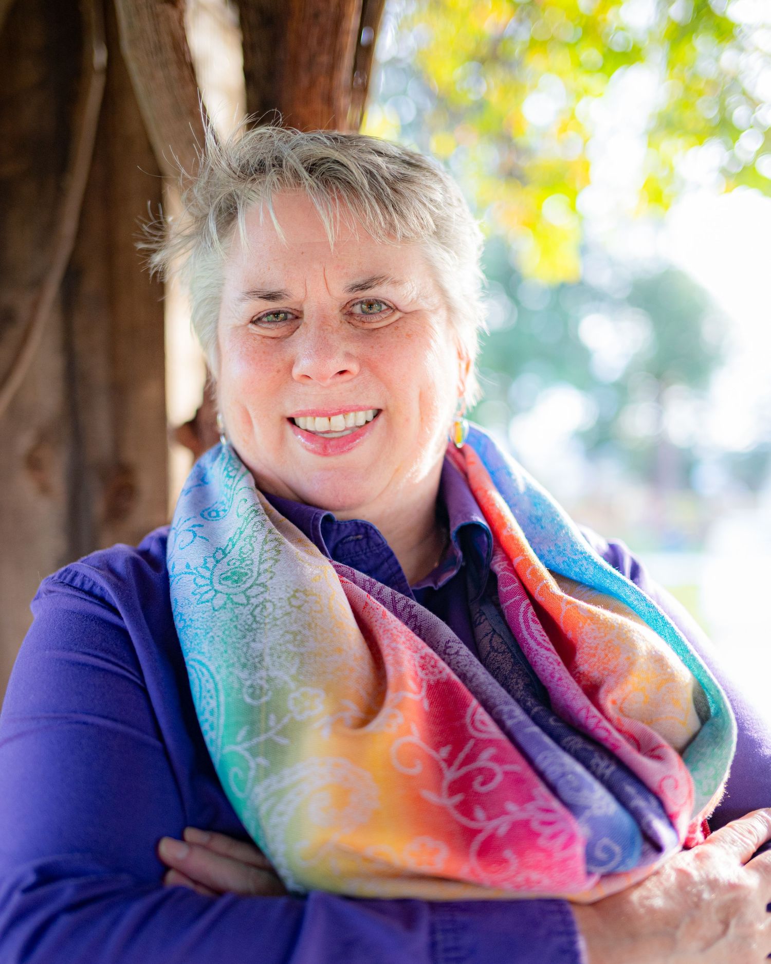 Portrait of someone wearing a vibrant rainbow scarf and purple top while smiling outdoors.